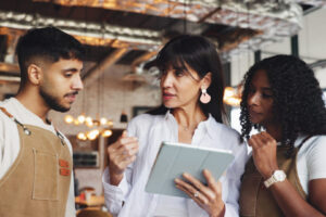 A woman wearing a white shirt and holding a tablet while standing between a man and a woman wearing brown aprons.