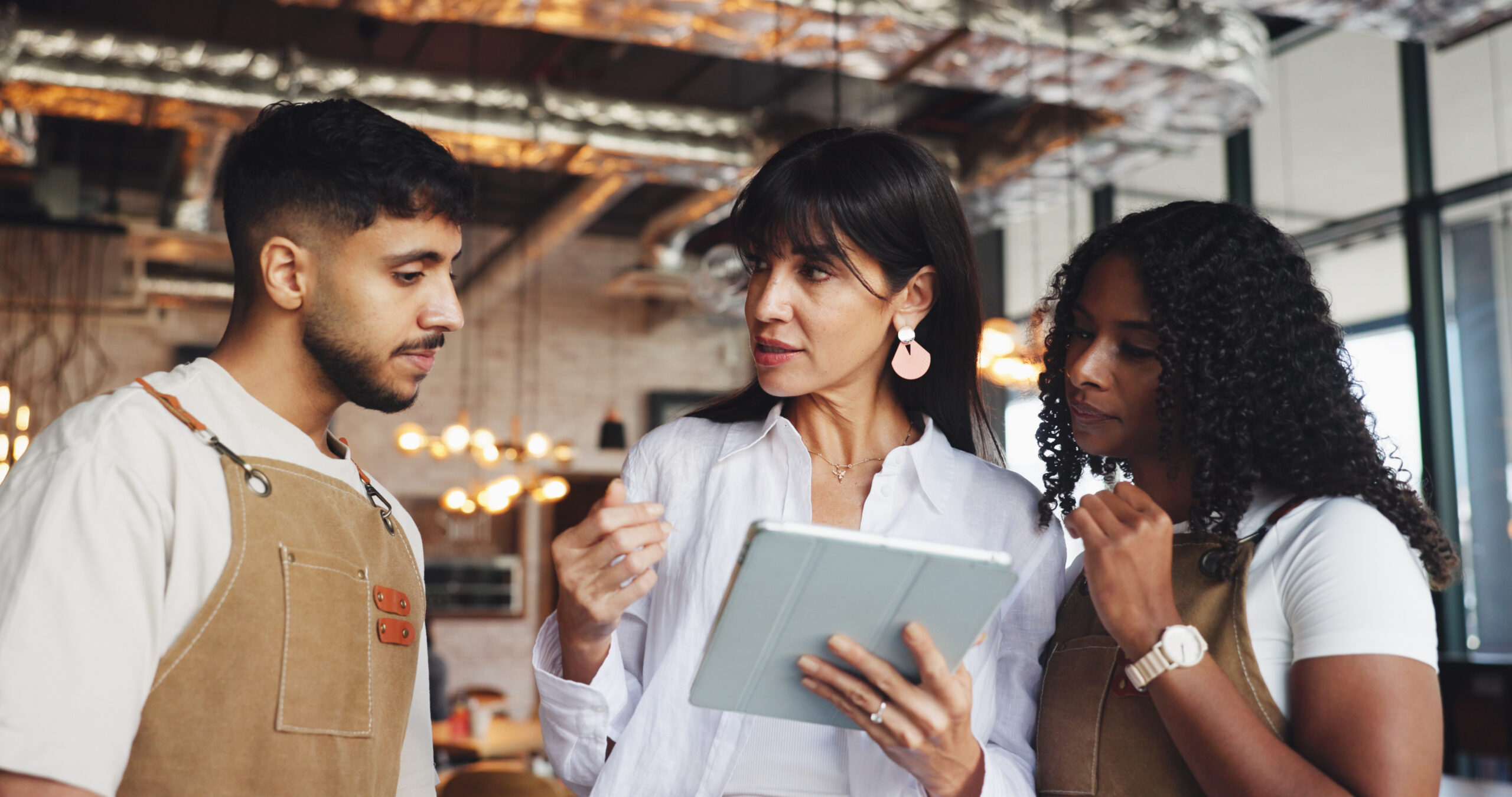 A woman wearing a white shirt and holding a tablet while standing between a man and a woman wearing brown aprons.