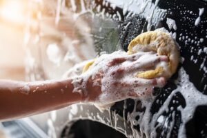 A person's hand is cleaning a car with a sponge. The car and the person's hand are covered in soap.