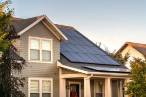 A two-story home with multiple rows of solar panels on the side roof surrounded by trees on a sunny day.