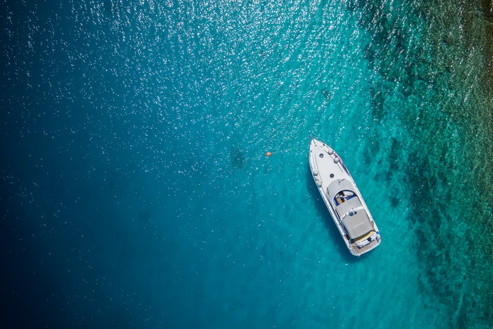 An aerial view of a white yacht floating over crystal clear water with a reef and rocks underwater to the right.