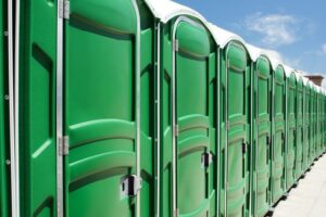 A row of green portable toilets lines a paved surface under a clear blue sky, extending into the distance in bright sunlight.