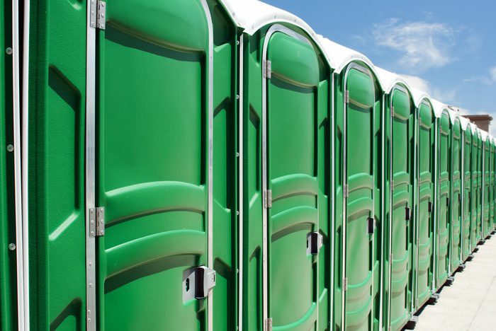 A row of green portable toilets lines a paved surface under a clear blue sky, extending into the distance in bright sunlight.