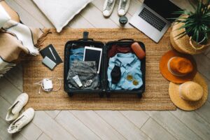 An open suitcase contains folded clothes, a tablet, and accessories, surrounded by shoes, a laptop, and other travel items.