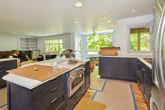 A kitchen with wood flooring covered in flat cardboard boxes, appliances covered in plastic and trees in the background.