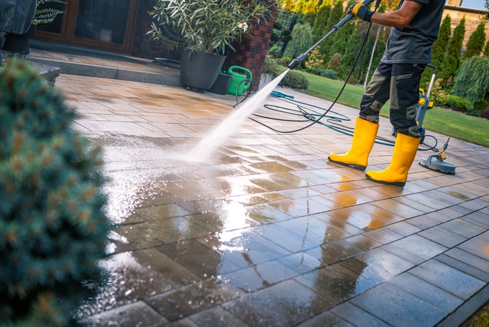 A worker in yellow boots sprays water across glossy pavers to pressure washes a stone patio beside a home.