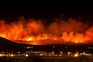 A wildfire burns across a dark ridgeline as orange smoke towers above buildings and lights in the valley below.