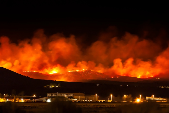 A wildfire burns across a dark ridgeline as orange smoke towers above buildings and lights in the valley below.